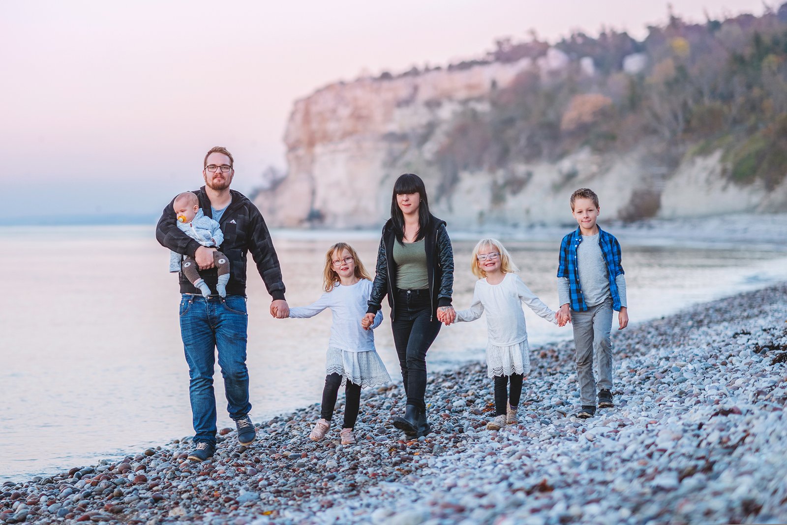 Familj går hand i hand längs den steniga stranden vid havet i kvällsljus, Fridhem.