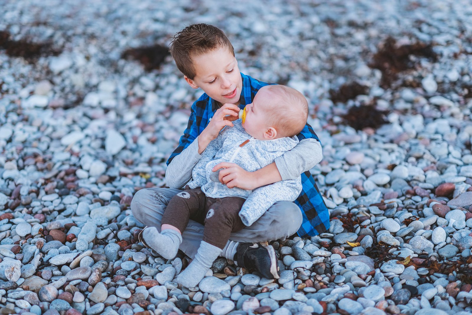 Två syskon sitter på den steniga stranden vid havet i kvällsljus.