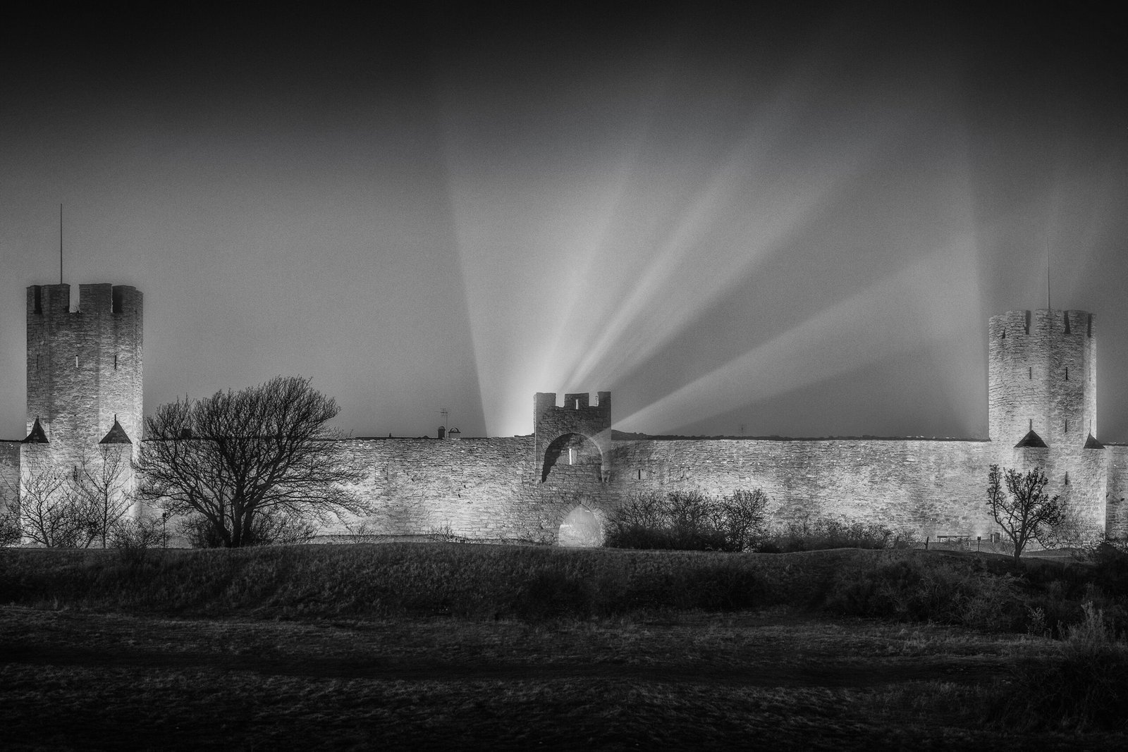 Svartvit bild av Visby ringmur en dimmig kväll med ljusstrålar som lyser genom dimman.